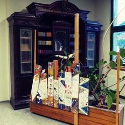 A room with a dark wooden cabinet, glass doors, and shelves filled with books. A potted plant with green leaves is in front of a window. A colorful display with cards is on a wooden stand.