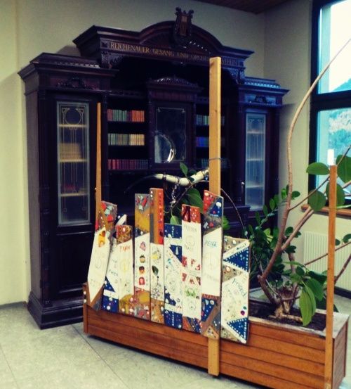 Ein Raum mit einem dunklen Holzschrank, Glastüren und Regalen voller Bücher. Ein Topf mit grünen Blättern steht vor einem Fenster. Eine bunte Ausstellung mit Karten steht auf einem Holzständer.