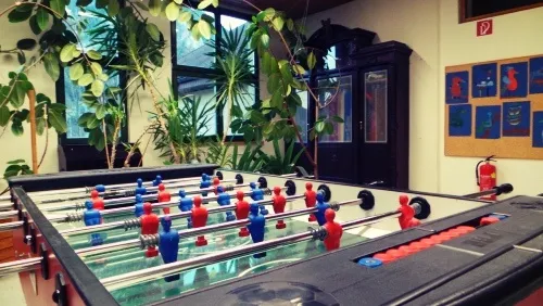 Ein Tischfußballtisch mit roten und blauen Spielern steht in einem Raum mit Schrank, Topfpflanzen und Feuerlöscher.
