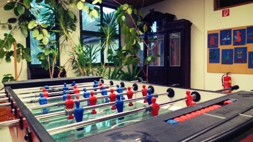Ein Tischfußballtisch mit roten und blauen Spielern steht in einem Raum mit Schrank, Topfpflanzen und Feuerlöscher.