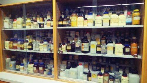 Ein hölzerner Schrank gefüllt mit zahlreichen Flaschen, Gläsern und Behältern. Die Flaschen haben verschiedene Farben und Größen, und einige haben Etiketten. Der Schrank hat Glastüren.