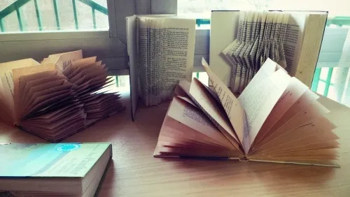 Ein Holztisch trägt mehrere Bücher. Einige sind in einzigartigen Origami-Stilen gefaltet, während andere offen sind. Ein Fenster mit einem grünen Geländer ist im Hintergrund.