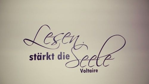 Ein weißer Hintergrund mit einem violetten kursiven Zitat, das 'Lesen stärkt die Seele' von Voltaire besagt.