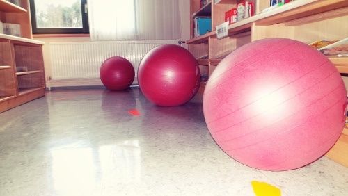 Ein Raum mit drei roten Übungsbällen auf dem Boden und einem Regal mit Gegenständen im Hintergrund.