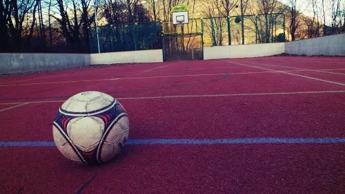 Ein Fußball liegt auf einer roten Laufbahn in einem Außensportplatz. Dahinter befindet sich ein Basketballkorb. Bäume umgeben das Gelände.