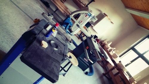 Eine Werkstatt mit einer Tischkreissäge, einem Hobel und anderen Holzbearbeitungswerkzeugen. Der Raum hat eine weiße Wand und eine Holzdecke.