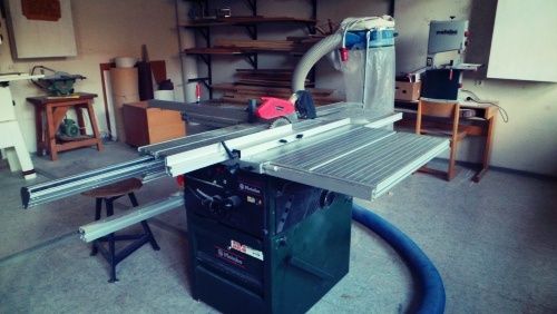 Eine Tischkreissäge steht in der Mitte einer Werkstatt. Dahinter befinden sich Regale mit Holz und anderen Geräten. Ein Hocker steht auf der linken Seite und ein blauer Schlauch liegt auf dem Boden.