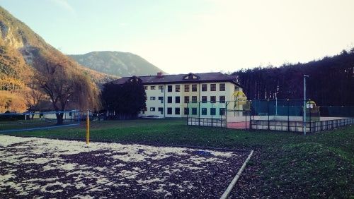 Ein großes Gebäude mit vielen Fenstern und einem Spielplatz befindet sich in einem grasbewachsenen Bereich mit einem Zaun. Im Hintergrund sind Berge zu sehen.