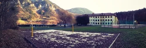Ein Außensportplatz mit einem Gebäude im Hintergrund, umgeben von Bergen und Bäumen. Der Platz hat ein Netz und einen Zaun.
