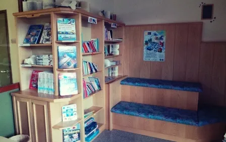 Ein kleiner Raum mit Holzwänden, Regalen voller Bücher und einer Holzbank mit blauen Kissen. Ein gerahmtes Bild ist an der Wand.