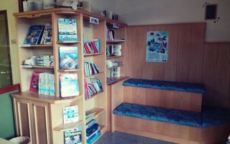 Ein kleiner Raum mit Holzwänden, Regalen voller Bücher und einer Holzbank mit blauen Kissen. Ein gerahmtes Bild ist an der Wand.