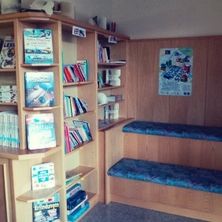 Ein kleiner Raum mit Holzwänden, Regalen voller Bücher und einer Holzbank mit blauen Kissen. Ein gerahmtes Bild ist an der Wand.