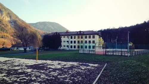 Ein Außensportplatz mit einem gelben Volleyballnetz vor einem großen Gebäude mit Bergen im Hintergrund.
