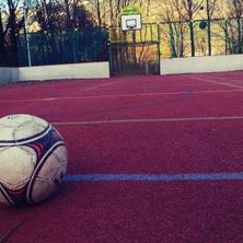 Ein Fußball liegt auf einem roten Feld mit weißen Markierungen, umgeben von einer Betonmauer. Im Hintergrund ist ein Basketballkorb zu sehen.