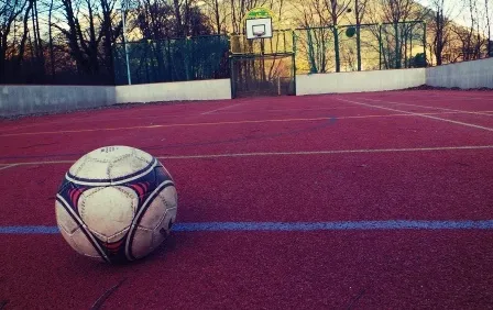 Ein Fußball liegt auf einem roten Feld mit weißen Markierungen, umgeben von einer Betonmauer. Im Hintergrund ist ein Basketballkorb zu sehen.