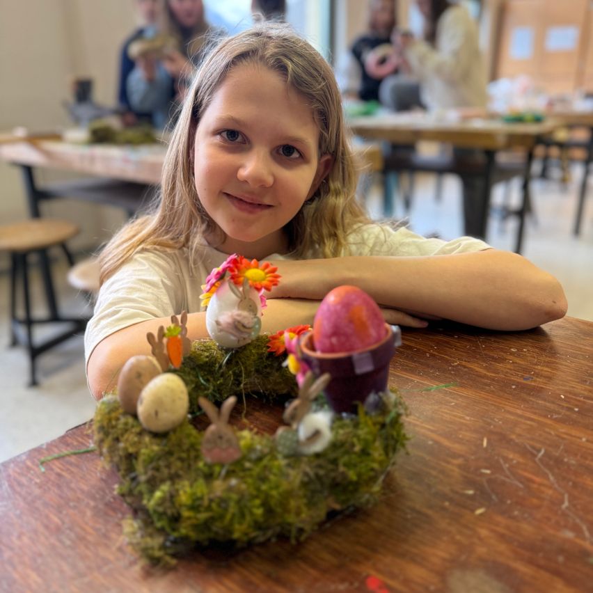 Ein Mädchen lächelt, während es an einem Tisch sitzt, umgeben von einem selbstgemachten Osterkranz mit Moos, Blumen und Ostereiern.