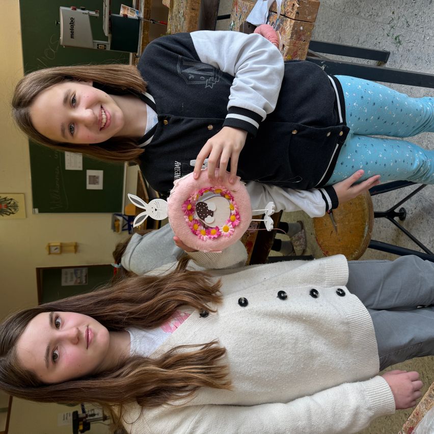 Zwei Mädchen posieren mit einem rosa Kuchen mit Blumen darauf. Ein Mädchen steht hinter dem anderen und hält den Kuchen.