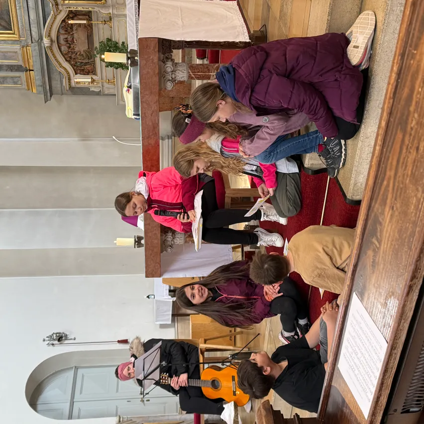 Gruppe von Kindern und Erwachsenen sitzen und stehen auf dem Boden einer Kirche, einige spielen Gitarre, einer spielt Trompete und ein anderer liest Noten.