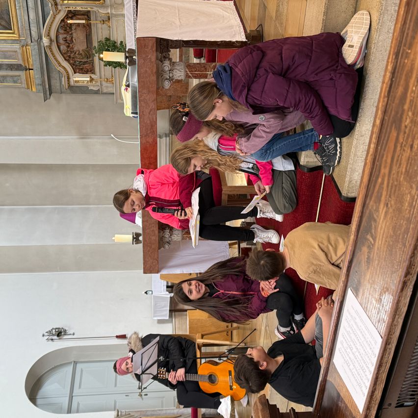 Gruppe von Kindern und Erwachsenen sitzen und stehen auf dem Boden einer Kirche, einige spielen Gitarre, einer spielt Trompete und ein anderer liest Noten.