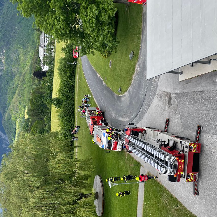 Ein Feuerwehrauto steht am Straßenrand, umringt von Menschen. Die Straße ist von Gras und Bäumen umgeben.
