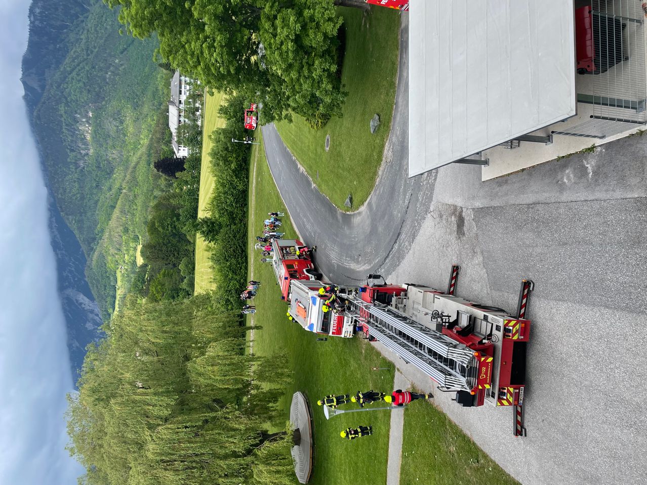 Ein Feuerwehrauto steht am Straßenrand, umringt von Menschen. Die Straße ist von Gras und Bäumen umgeben.