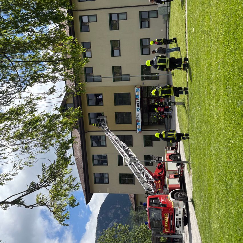 Feuerwehrleute stehen vor einem Gebäude mit einem Feuerwehrauto, einer Leiter und einer Person auf der Leiter. Das Gebäude hat viele Fenster und ist von Gras umgeben.