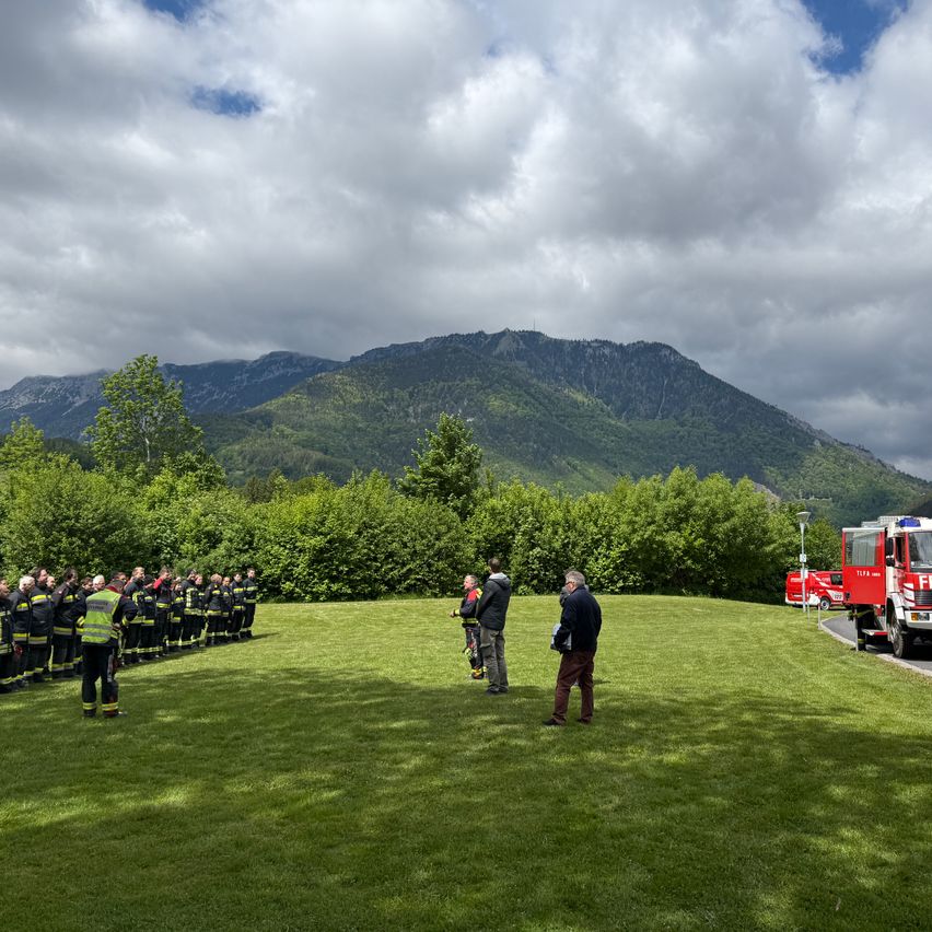 Eine Gruppe von Feuerwehrleuten in Uniform steht auf einem Rasen mit Bergen im Hintergrund. Ein Mann spricht zu ihnen.