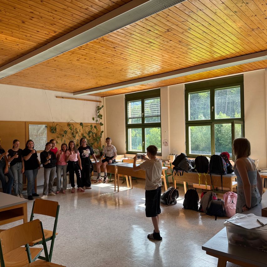 Ein Klassenzimmer mit Holzdecke und Fenstern. Mehrere Schüler sind versammelt, einige machen Fotos mit Kameras. Rucksäcke sind auf Bänken und Tischen verstreut.