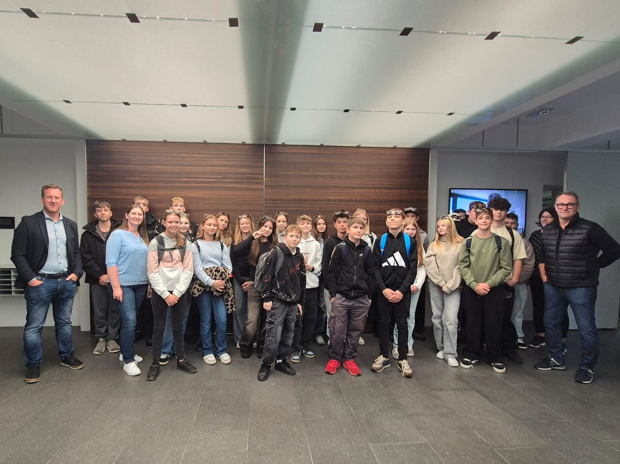 Eine Gruppe junger Menschen posiert für ein Foto in einem Raum mit Holzwand und weißer Decke. Einige tragen Hüte und Rucksäcke.