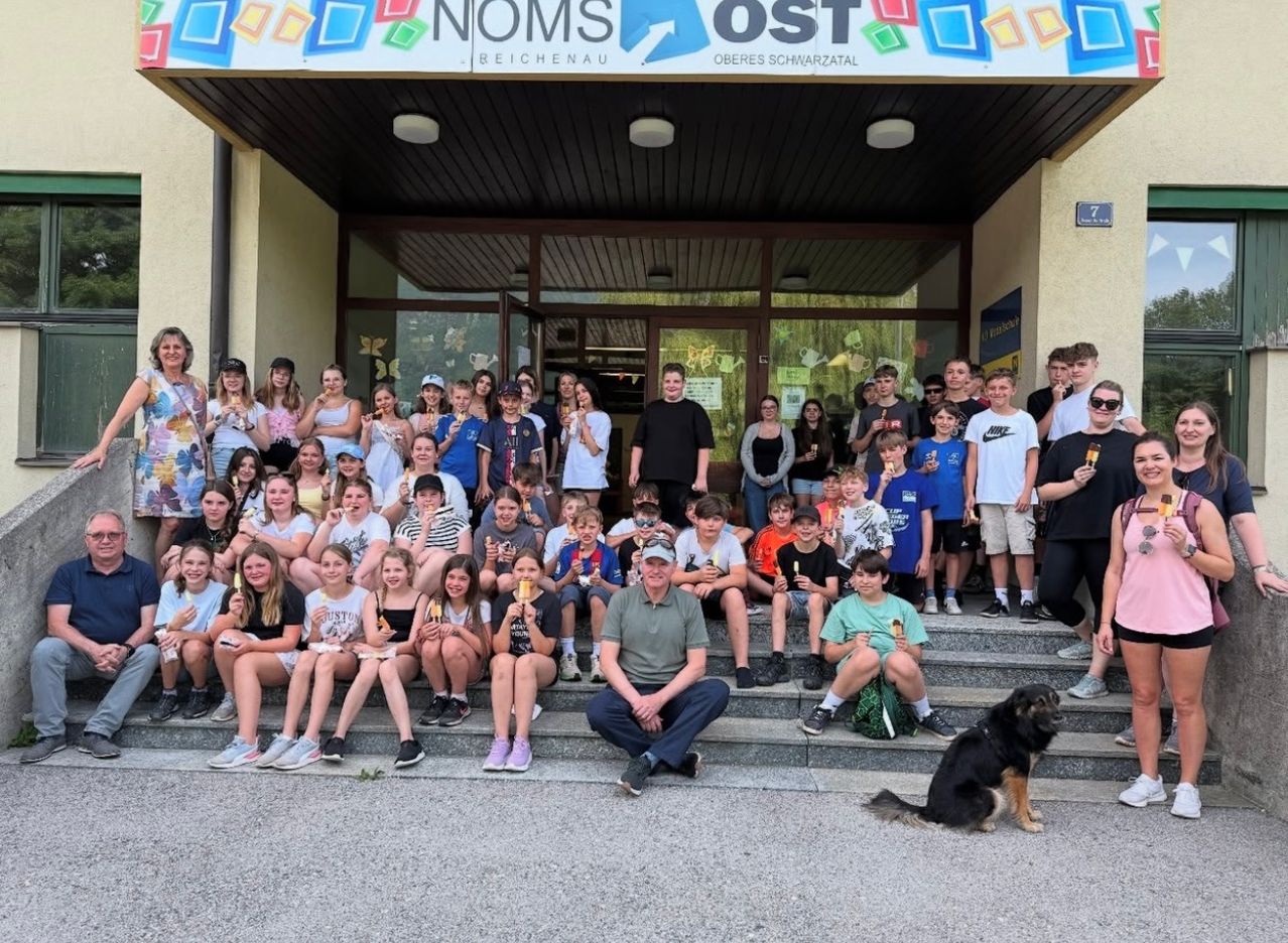 Eine Gruppe von Menschen, wahrscheinlich Schüler, versammelt sich vor einem Gebäude mit einem Banner, auf dem 'NOMS OST' steht. Sie halten Eis und posieren für ein Foto. Ein Mann sitzt auf den Stufen, und ein Hund sitzt neben ihm. Das Gebäude hat Glasfenster und eine Glastür.
