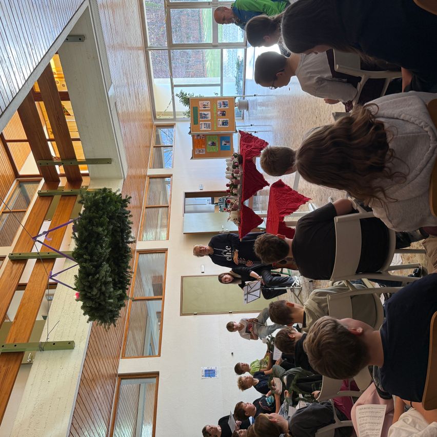 Eine Gruppe von Schülern versammelt sich für eine musikalische Aufführung, wobei eine Person im Vordergrund ein Instrument spielt, während andere auf Stühlen sitzen und zuschauen. Ein Weihnachtsbaum hängt oben und Poster sind an der Wand angebracht.