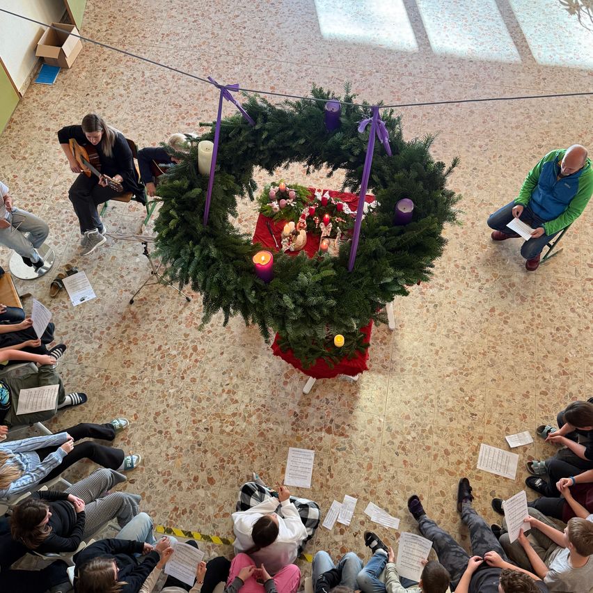 Eine Gruppe von Menschen sitzt um einen kreisförmigen Adventskranz mit Kerzen herum und hält Papiere und Instrumente. Eine Person spielt Gitarre, während andere beobachten.