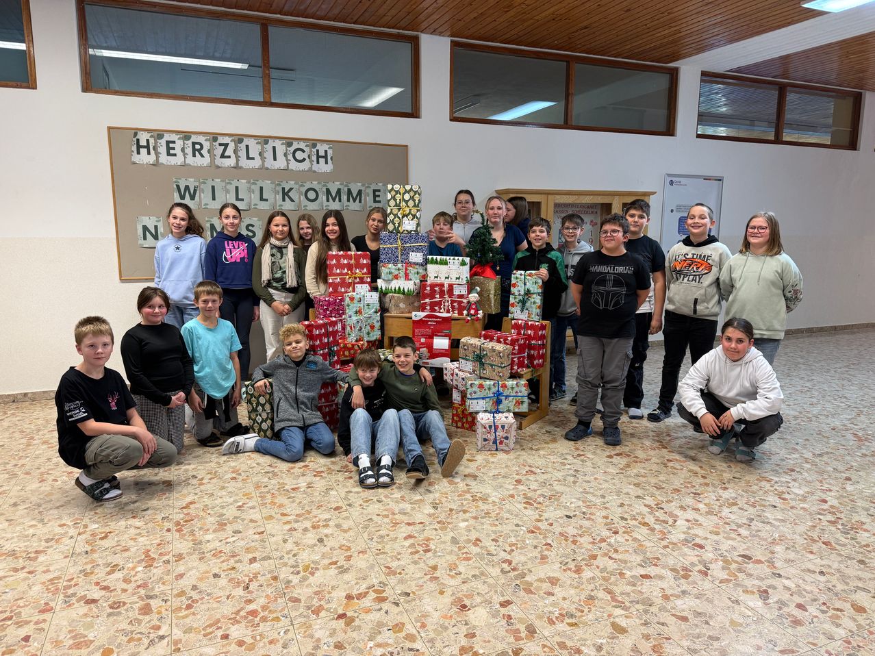 Eine Gruppe von Kindern und Jugendlichen posiert für ein Foto mit vielen verpackten Geschenken auf dem Boden. Der Raum hat eine Holzdecke und große Fenster. Die Wand hat eine Nachricht auf Deutsch.