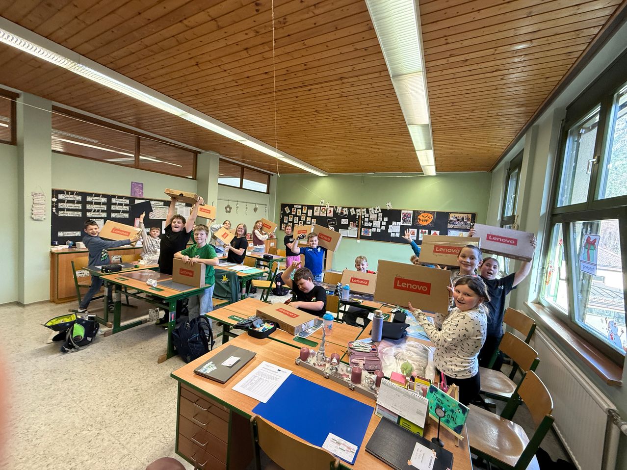 Ein Klassenzimmer voller Schüler, die Lenovo-Boxen hochhalten. Einige sitzen an Schreibtischen, andere stehen. Helle Beleuchtung und Poster schmücken den Raum.