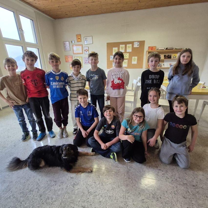 Eine Gruppe von Kindern und ein Hund posieren für ein Foto in einem Klassenzimmer mit Tischen, Stühlen und einer Pinnwand.