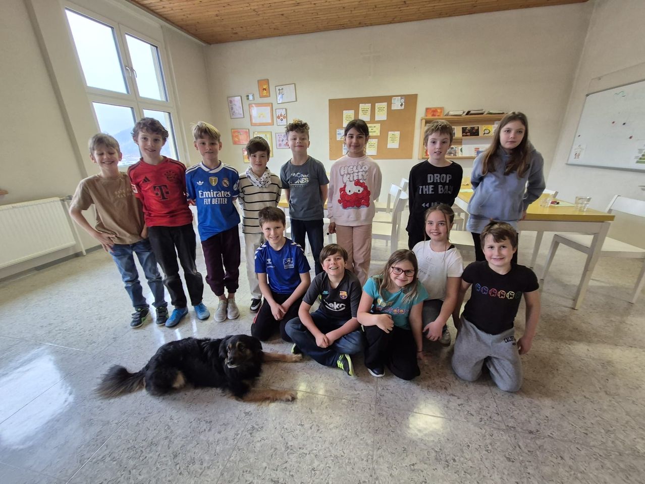 Eine Gruppe von Kindern und ein Hund posieren für ein Foto in einem Klassenzimmer mit Tischen, Stühlen und einer Pinnwand.