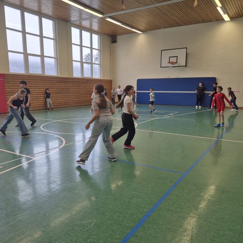 Mehrere Kinder spielen Basketball in einer Turnhalle. Ein Mädchen geht zum Korb, und ein anderes folgt. Ein Basketballkorb ist an der Wand.