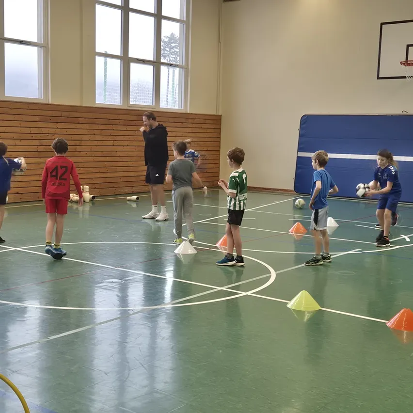 Eine Gruppe von Kindern spielt drinnen Fußball. Ein Mann steht vor ihnen und scheint Anweisungen zu geben. Es gibt Kegel auf dem Boden und einige Kinder halten Fußballbälle.