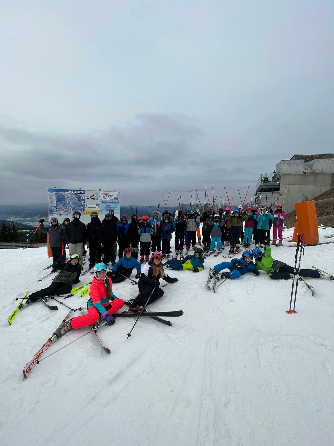 Eine Gruppe von Skifahrern und Snowboardern in bunter Winterkleidung posiert für ein Foto auf einem verschneiten Berg. Einige sitzen auf dem Boden, während andere hinter ihnen stehen.
