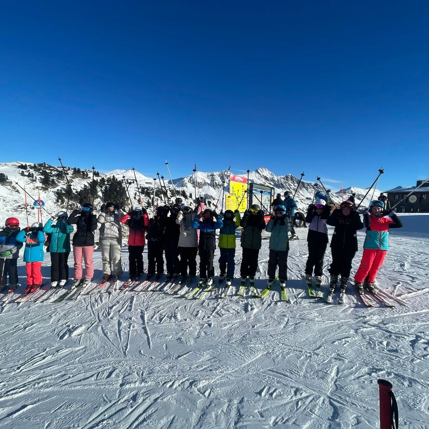 Eine Gruppe junger Skifahrer und Snowboarder posiert für ein Foto auf einem verschneiten Berg unter blauem Himmel.
