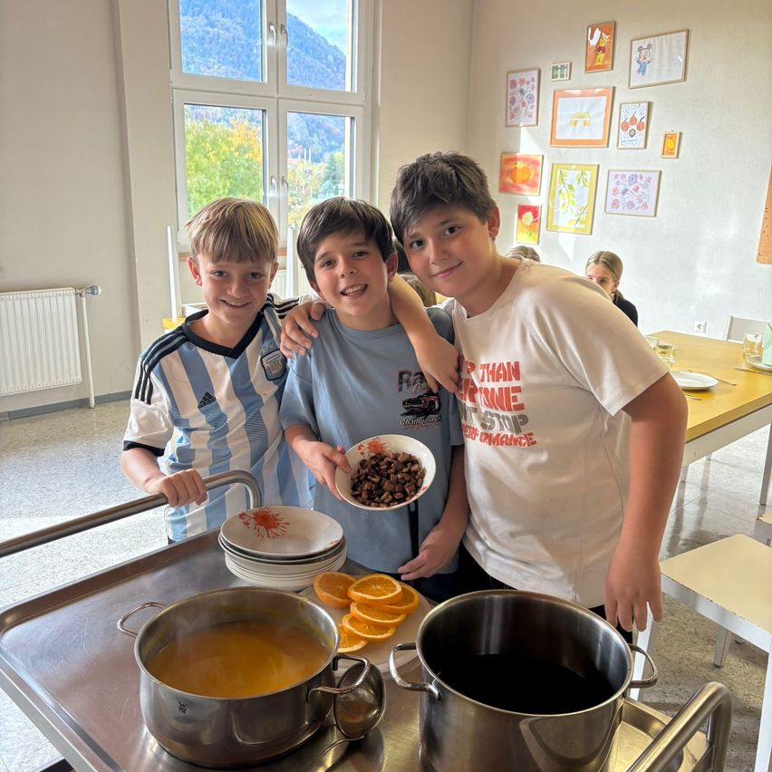 Drei Jungen stehen in einem Raum mit einem Kochaufbau. Sie halten Schalen mit Essen. Es gibt gestapelte Teller, Töpfe und Orangen. Ein Tisch und Stühle sind in der Nähe. Kunstgemälde hängen an der Wand.