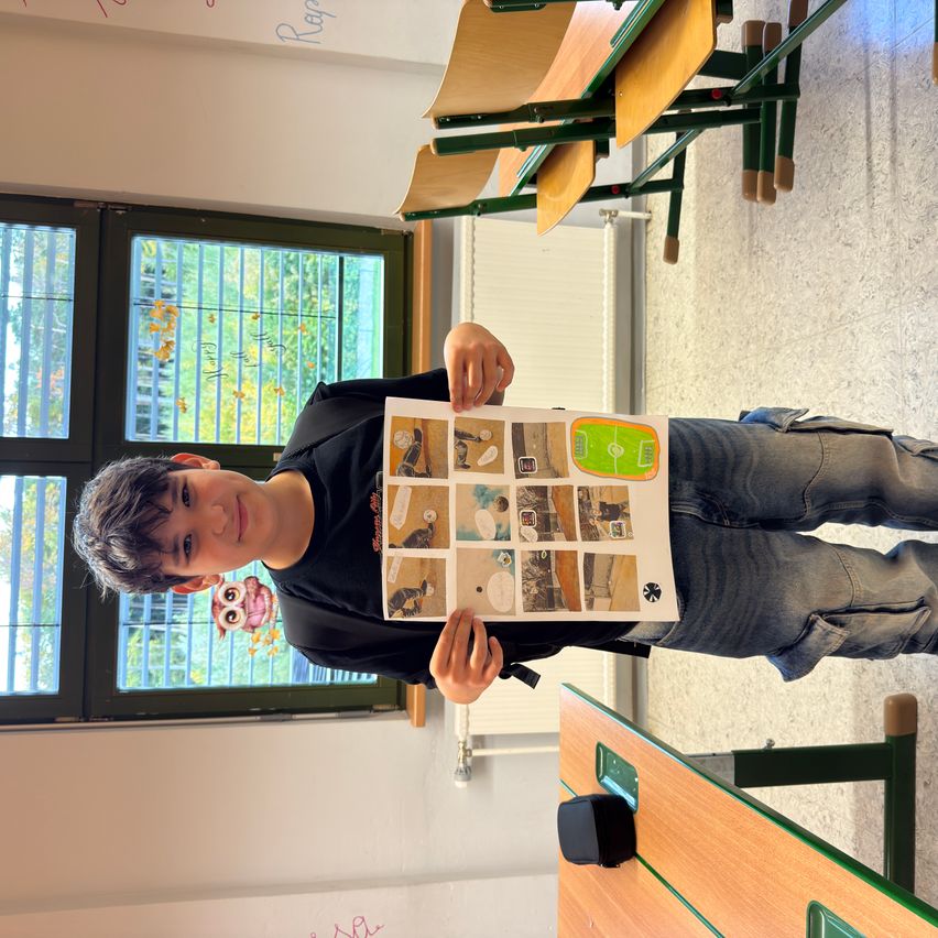 Ein Junge steht in einem Klassenzimmer, hält ein Stück Papier mit einem Bild darauf und lächelt. Hinter ihm sind Stühle gestapelt und ein Fenster mit Bäumen draußen.