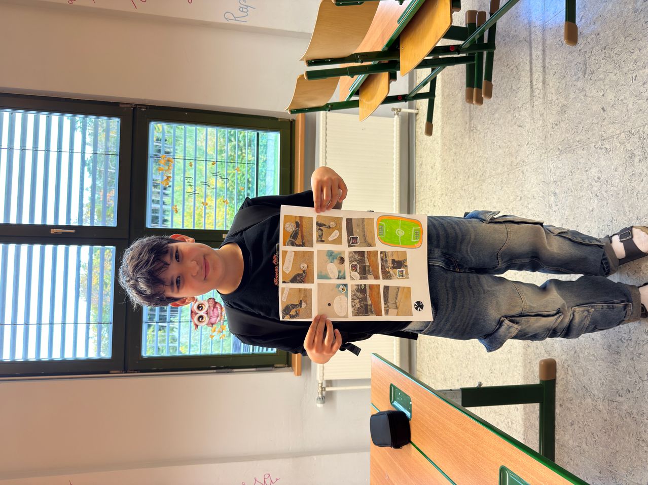 Ein Junge steht in einem Klassenzimmer, hält ein Stück Papier mit einem Bild darauf und lächelt. Hinter ihm sind Stühle gestapelt und ein Fenster mit Bäumen draußen.