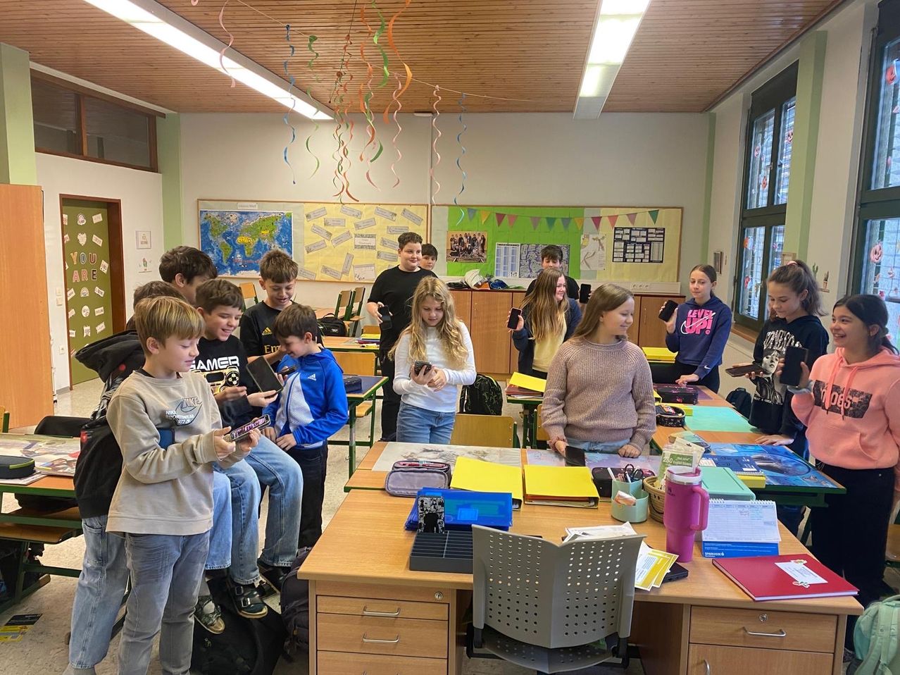 Ein Klassenzimmer voller Schüler, die ihre Handys benutzen; einige sitzen auf Tischen, während andere stehen, mit Dekorationen und Postern an den Wänden.