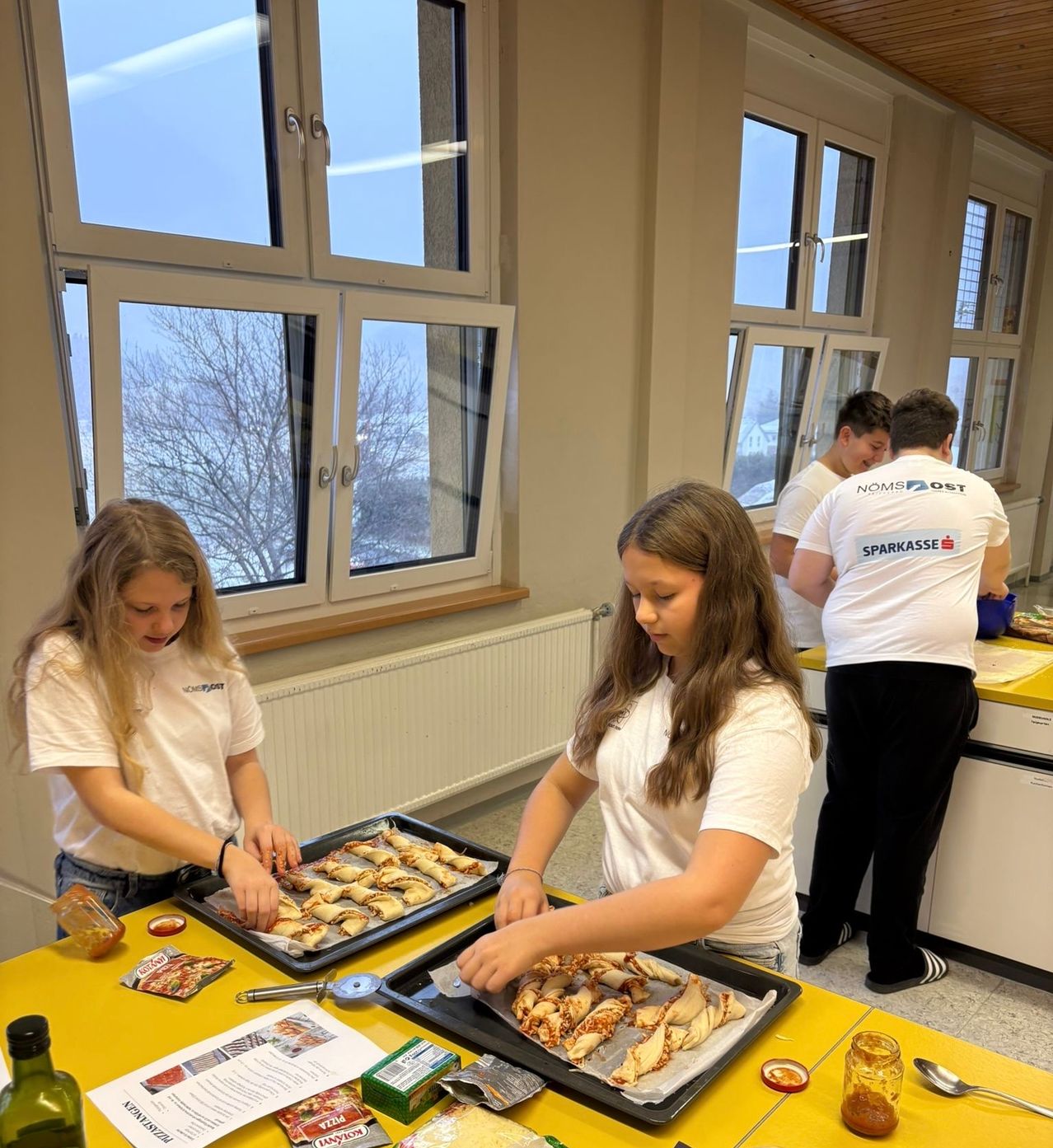 Zwei Mädchen bereiten auf einem gelben Tisch in einer Küche mit großen Fenstern Essen zu. Sie tragen weiße T-Shirts und scheinen Pizza zuzubereiten. Zwei Männer stehen hinter ihnen, ebenfalls in weißen T-Shirts. Es gibt einen Heizkörper und eine blaue Schüssel auf der Arbeitsplatte.