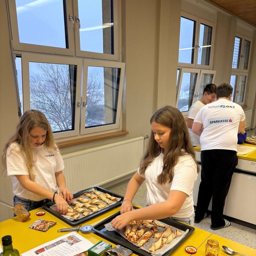 Zwei Mädchen bereiten auf einem gelben Tisch in einer Küche mit großen Fenstern Essen zu. Sie tragen weiße T-Shirts und scheinen Pizza zuzubereiten. Zwei Männer stehen hinter ihnen, ebenfalls in weißen T-Shirts. Es gibt einen Heizkörper und eine blaue Schüssel auf der Arbeitsplatte.