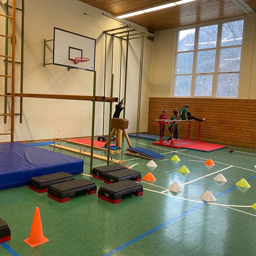 Ein Indoor-Fitnessstudio mit verschiedenen Trainingsgeräten, darunter ein Basketballkorb, eine Balancestange und Bodenmatten. Kinder sind bei Aktivitäten auf dem Boden des Fitnessstudios zu sehen.