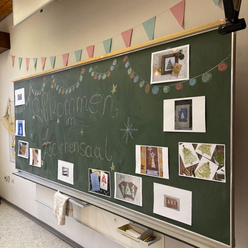 Ein Klassenzimmer-Schwarzbrett, das mit Feiertagsbildern und Bastelarbeiten dekoriert ist, einschließlich einer Girlande, die 'Willkommen im Weihnachtssaal' sagt.