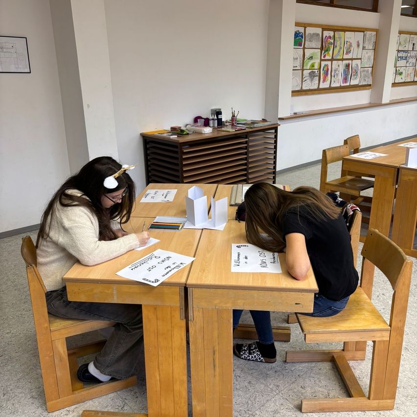 Zwei Mädchen sitzen an einem Holzschreibtisch, eine schreibt auf Papier und die andere ruht ihren Kopf auf ihren Armen. Sie befinden sich in einem Klassenzimmer mit Kunstwerken an der Wand und einem Holzschrank im Hintergrund.