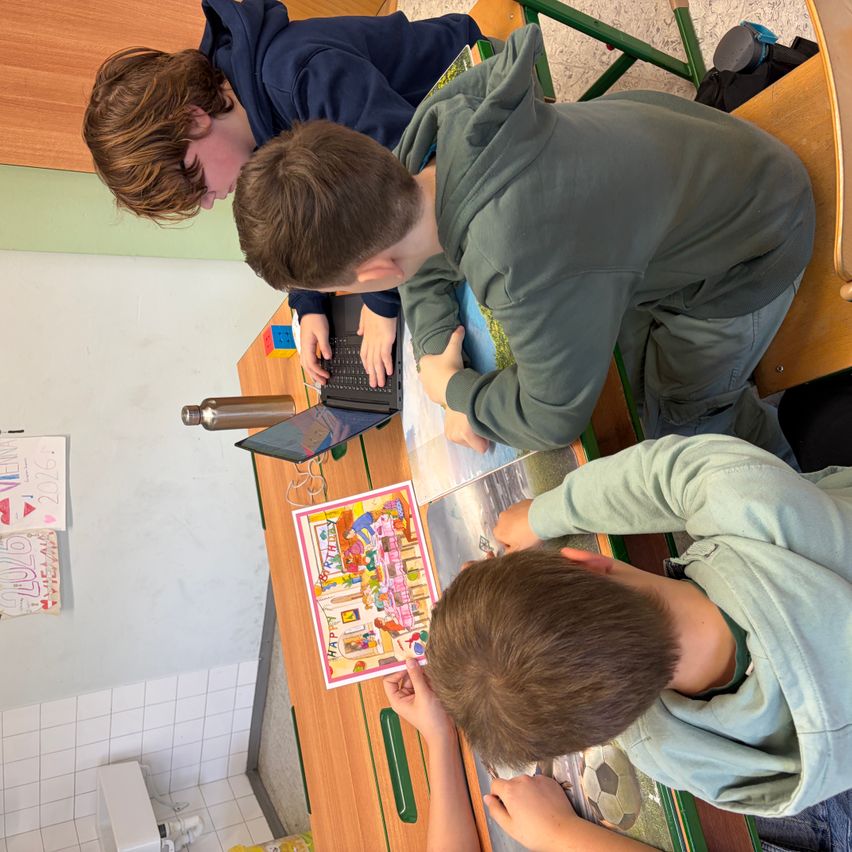 Vier Jungen arbeiten gemeinsam an einem Laptop und einem Malbuch an einem Schreibtisch im Klassenzimmer. Ein Junge tippt auf dem Laptop, während andere ein Malbuch betrachten.